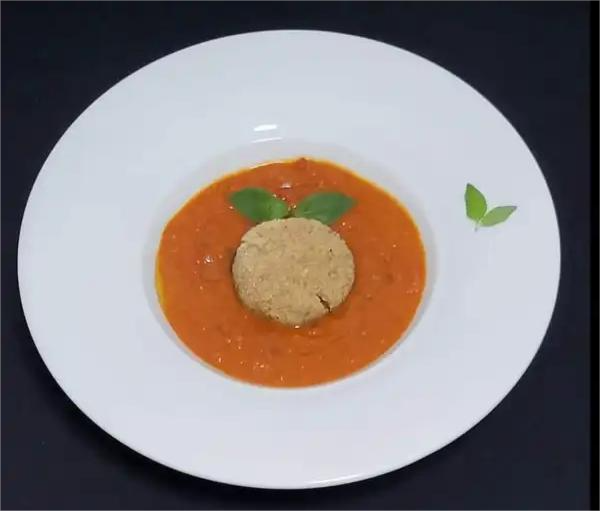 Un piatto di zuppa di pomodoro affumicato con una pralina di ricotta di soia al centro, guarnito con foglie verdi su fondo scuro.