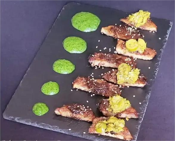 Piatto gourmet vegano con pez a fette, guarnito con salsa verde e pezzi di verdura, presentato su un vassoio nero.
