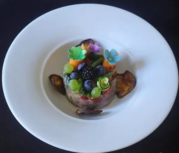 Un piatto di antipasto vegano guarnito con frutta, verdura e decorazioni floreali, presentato in un contenitore circolare su un piatto bianco.