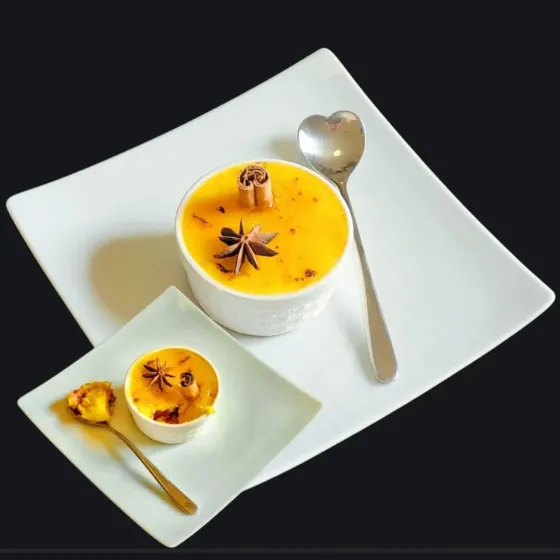 Un dessert vegano gourmet servito in una ciotola bianca decorata con una stecca di cannella e anice stellato, accanto a un piccolo piatto con una porzione del dessert e un cucchiaino dorato.