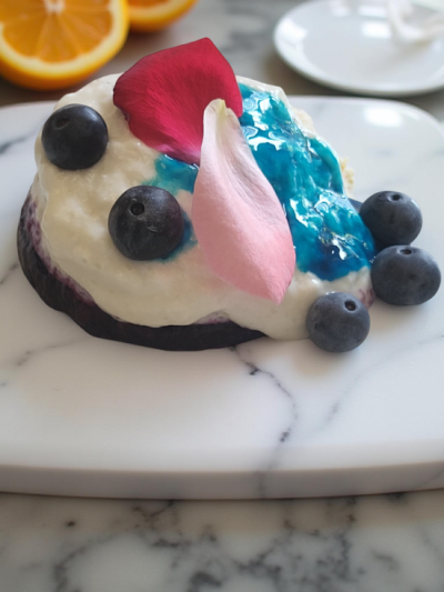 Dessert on a marble plate, featuring a creamy white topping, blue jelly, blueberries, and a pink petal for decoration.