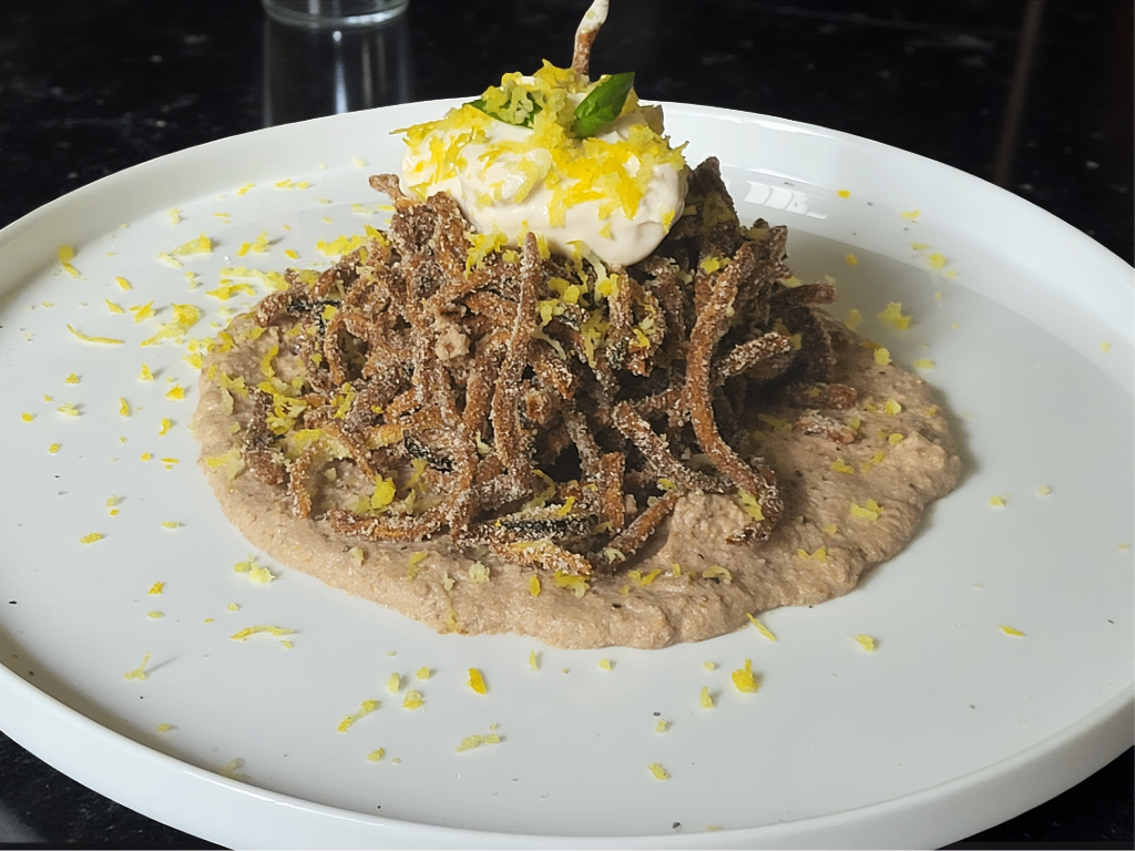 Piatto di menù vegano gourmet con noodles fritti, salsa cremosa e scorza di limone su un letto di crema di fagioli.