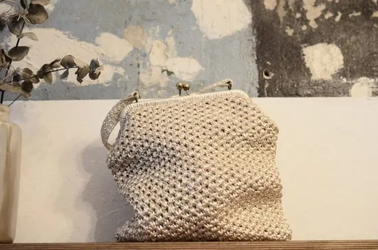 Una borsa in tessuto intrecciato di colore chiaro, appoggiata su una superficie di legno, con una decorazione di foglie secche sullo sfondo.