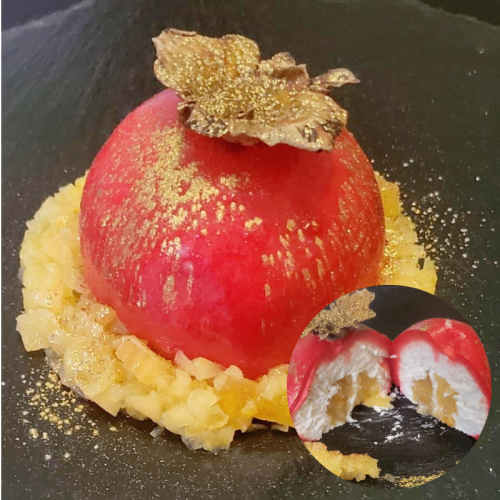 Dessert rotondo decorato con una foglia dorata in cima, circondato da cubetti di frutta candita, con un particolare che mostra l'interno cremoso e fruttato.
