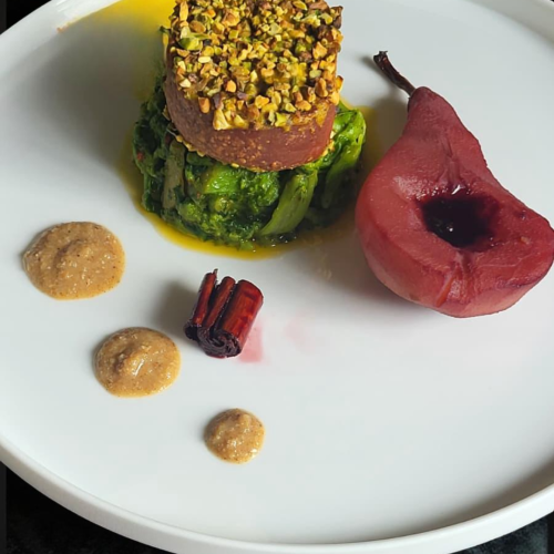 Piatto gourmet con una base di verdure verdi, un tortino ricoperto di pistacchi e una pera rossa glassata, accompagnato da salse di diverse consistenze.