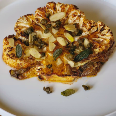 Una fetta di cavolfiore arrostito con mandorle, capperi e foglie di salvia su un piatto bianco.